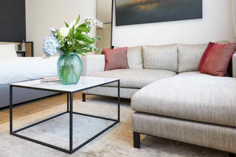 Citysuites Aparthotel Salford  Ngoại thất bức ảnh The photo features a modern living area. There is a light-colored, comfortable sectional sofa with decorative cushions in shades of red. A glass coffee table with a black metal frame is positioned in front of the sofa. On the table, there is a striki