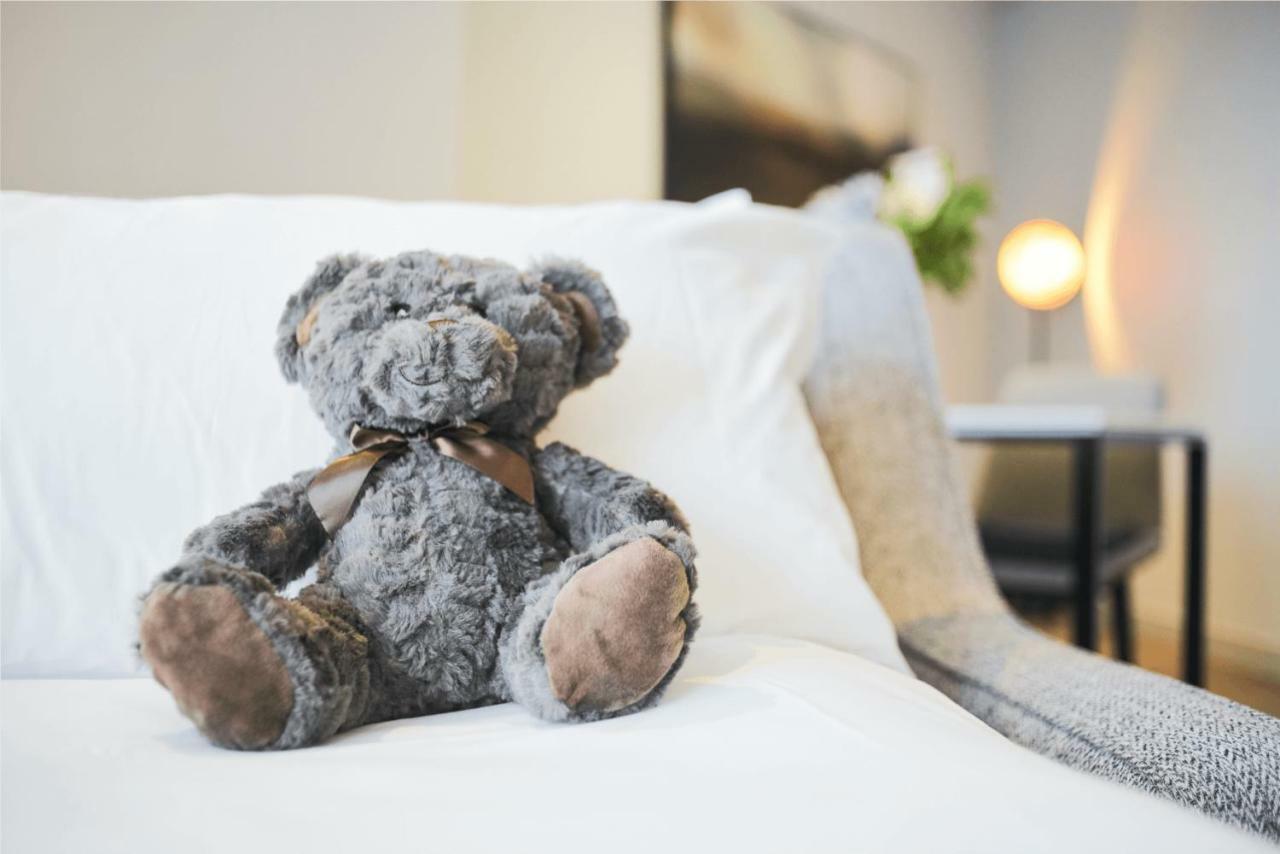 Citysuites Aparthotel Salford  Ngoại thất bức ảnh The photo shows a plush teddy bear, which is gray in color, sitting on a white couch. The bear has a brown ribbon tied around its neck and has seen some wear, with noticeable shading on its paws. In the background, there is a softly lit environment w