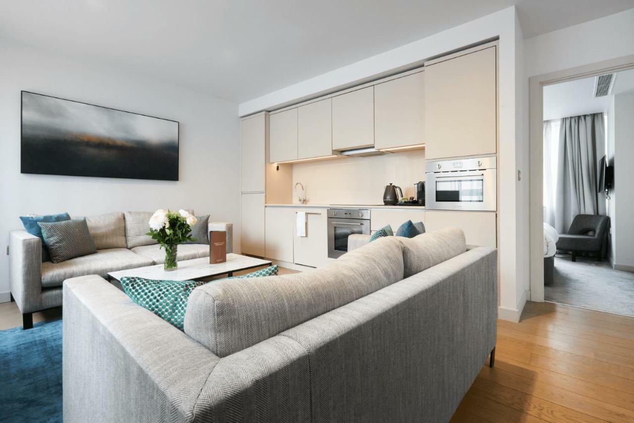 Citysuites Aparthotel Salford  Ngoại thất bức ảnh The photo shows a modern living space that includes a cozy living area and a small kitchen. The living area features a light gray sectional sofa with several pillows and a coffee table with a decorative centerpiece, possibly flowers. The kitchen, loc