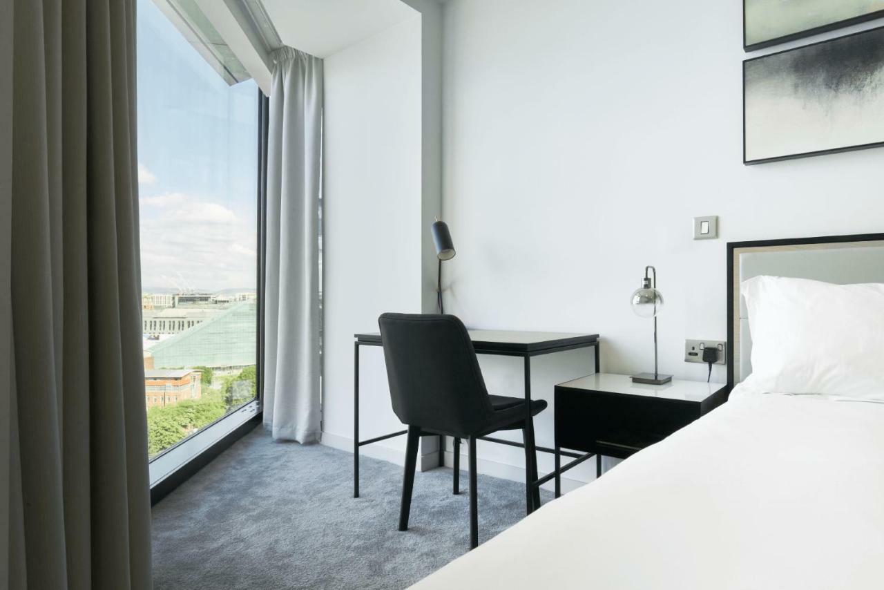 Citysuites Aparthotel Salford  Ngoại thất bức ảnh The photo shows a modern hotel room featuring a bed with crisp white linens. To one side, there is a large window with a view of the outside, framed by sheer curtains that allow natural light to enter. A sleek black desk and chair are positioned next